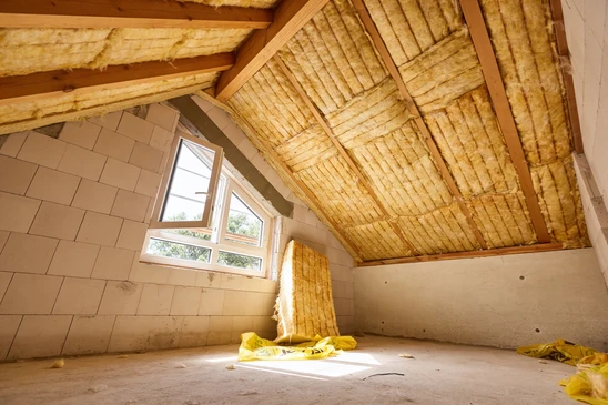 Ouvrier en combinaison de protection intégrale et masque respiratoire installant de l'isolant thermique jaune dans les combles d'une habitation